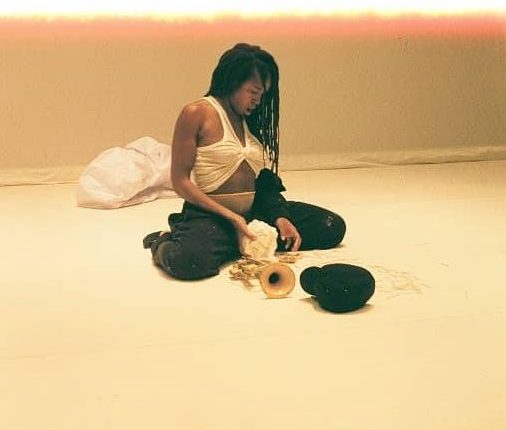 Dana Michel is sitting on the grown, her feet tucked under her body and her knees pointing outwards, wearing a white sleeveless shirt with the front tucked up in the colour and dark pants. There are a number of objects including a dark, unclear object and a trumpet in front of her and she holds a white unclear object. Behind Michel is a white tarp. The whole image has a warm tone to it, and the top border of the image is seems burnt out as if from an over exposure. The top border is irregular with a rough edge that turns orange and then sharply white.