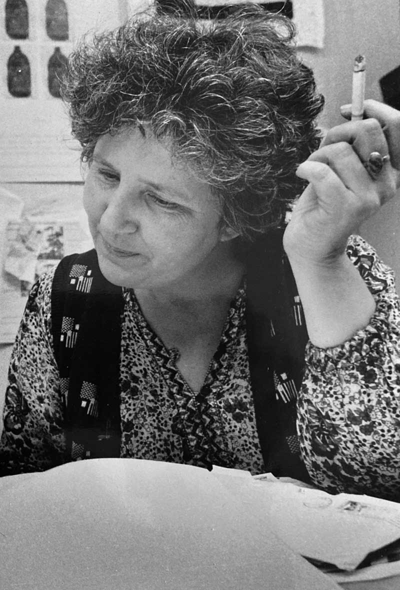 A black and white photograph of Ann Rosenberg smiling and looking at a book on a desk, with a cigarette in her hand. 