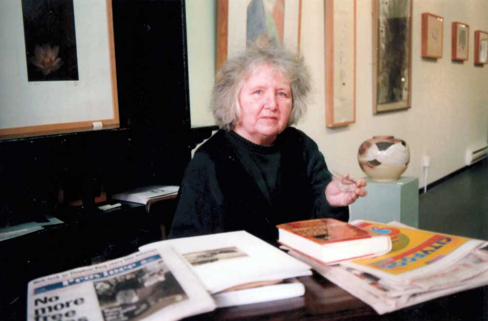 A colour photograph of Ann Rosenberg sitting in a gallery-like space behind a table full of newspapers and books. Framed artworks hang in the background.