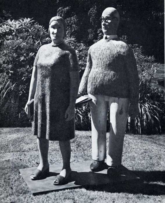 A greyscale photograph documenting two life-size figurative scluptures. A man wears a shirt, sweater, trousers, and glasses and holds a book; a woman wears a short-sleeved dress, sandals, and also holds a book.