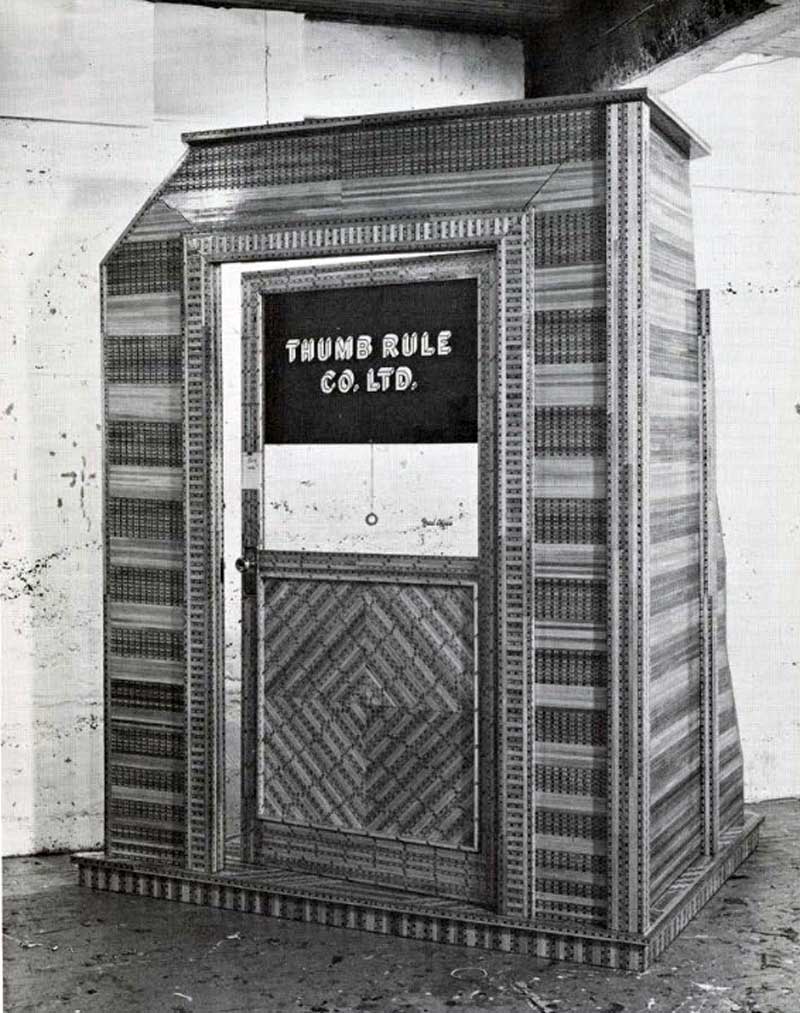 A greyscale image of Gary Lee Nova and Alan Miller's "Out to Metric" installation, depicting a lifesize kiosk made up of sections of wooden rulers. A sign on the door's black, roll-down blind reads "Thumb Rule Co. Ltd."