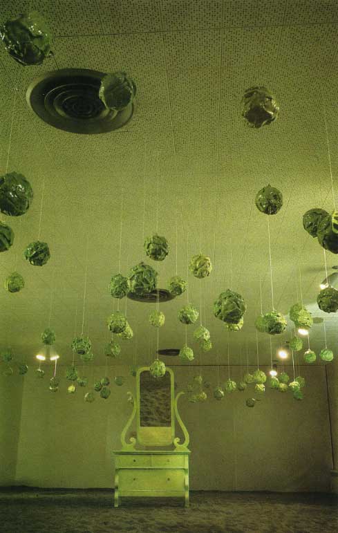 A green-lit installation image of Gathie Falk's "150 Cabbages" showing dozens of ceramic cabbage sculptures suspended from string from the ceiling, and an antique dresser and mirror in the distance. 