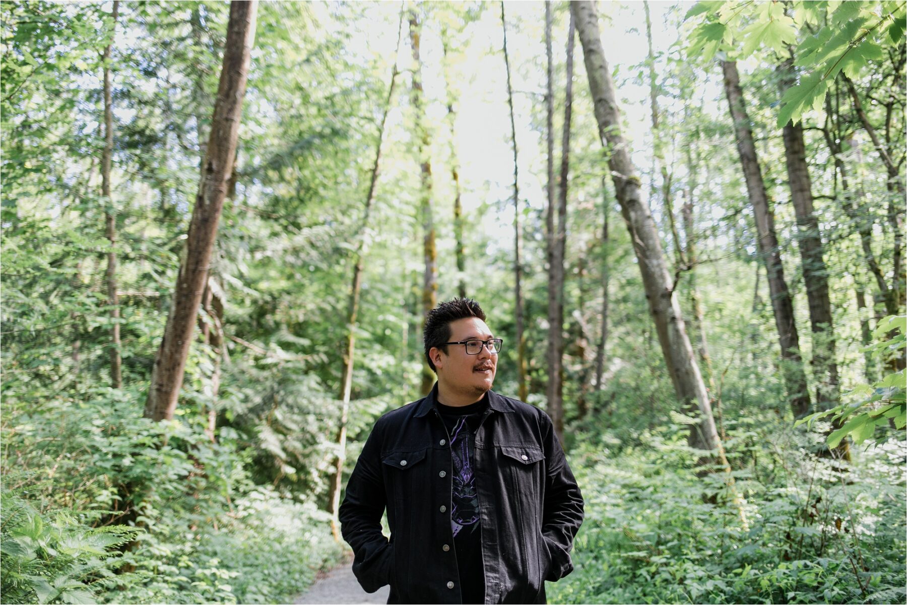 Photo of the author Jordan Abel standing in a forest and wearing glasses and a black jacket.