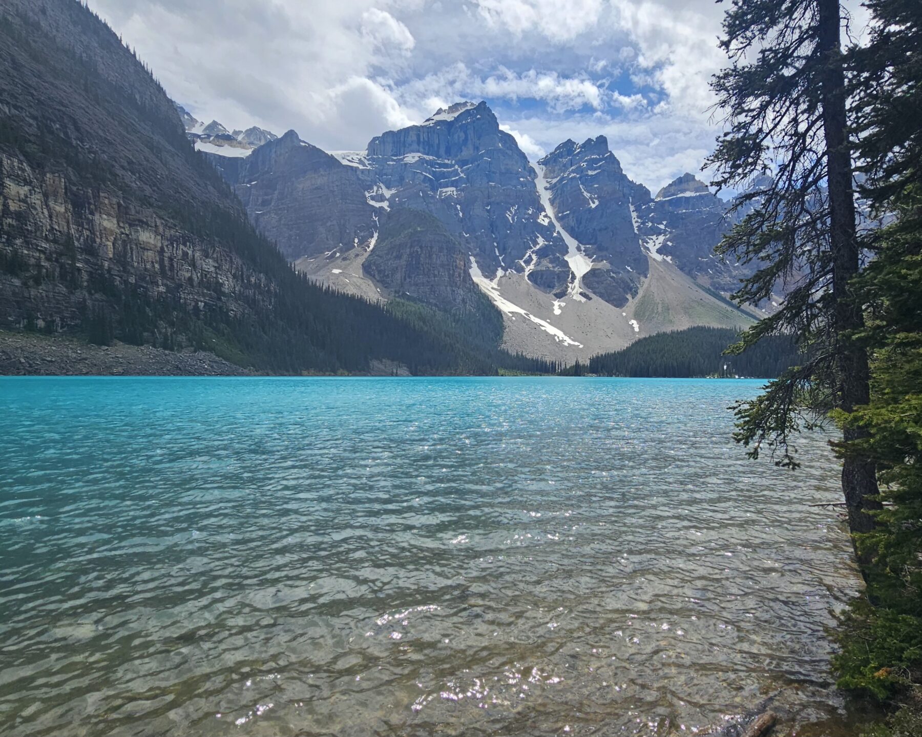 Photograph looking out at the jagged, snow-capped mountains from a sunny, turquoise lake framed by evergreen trees.